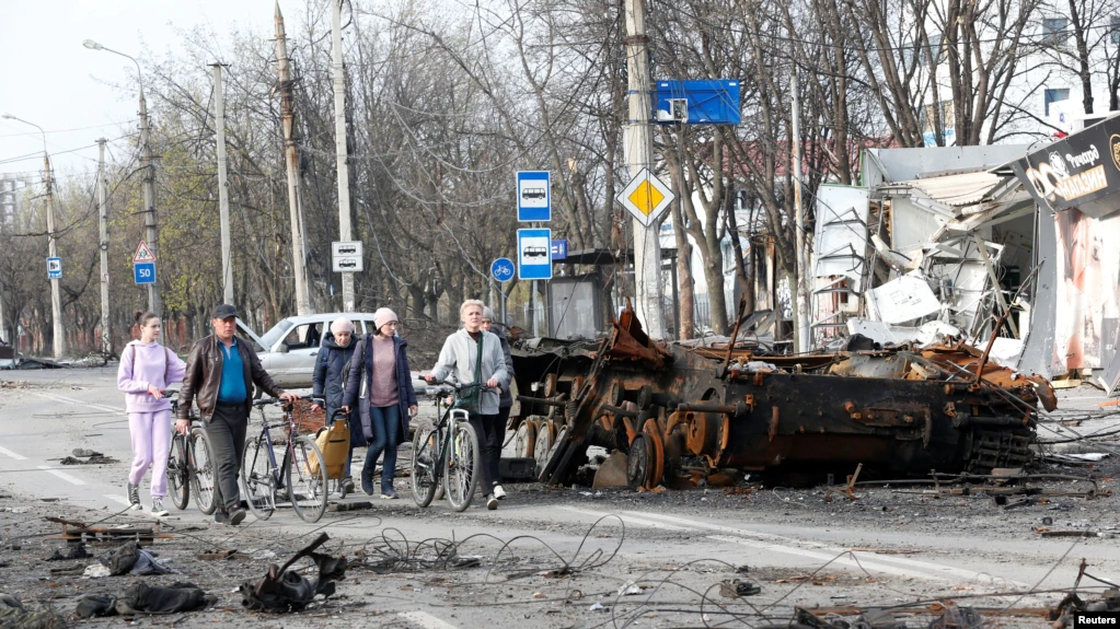 Oameni trecând pe lângă un tanc distrus în timpul conflictului dintre Ucraina și Rusia în orașul-port din sudul Ucrainei, Mariupol, 17 aprilie 2022